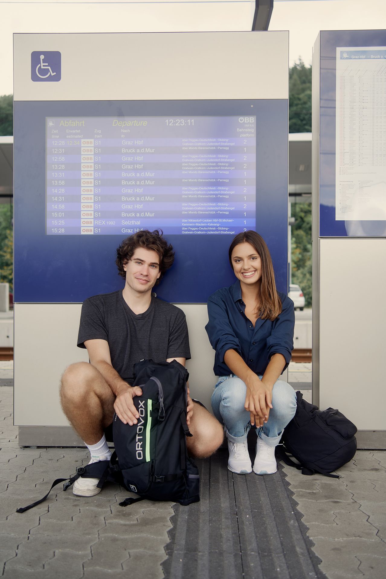Zwei Personen sitzen an einem Bahnhof. Die Frau trägt ein blaues Hemd und Jeans, der Mann ein graues T-Shirt. Sie lächeln beide und sitzen mit gekreuzten Beinen da, mit Rucksäcken. Dahinter zeigt ein digitales Board Abfahrtszeiten und -orte an.