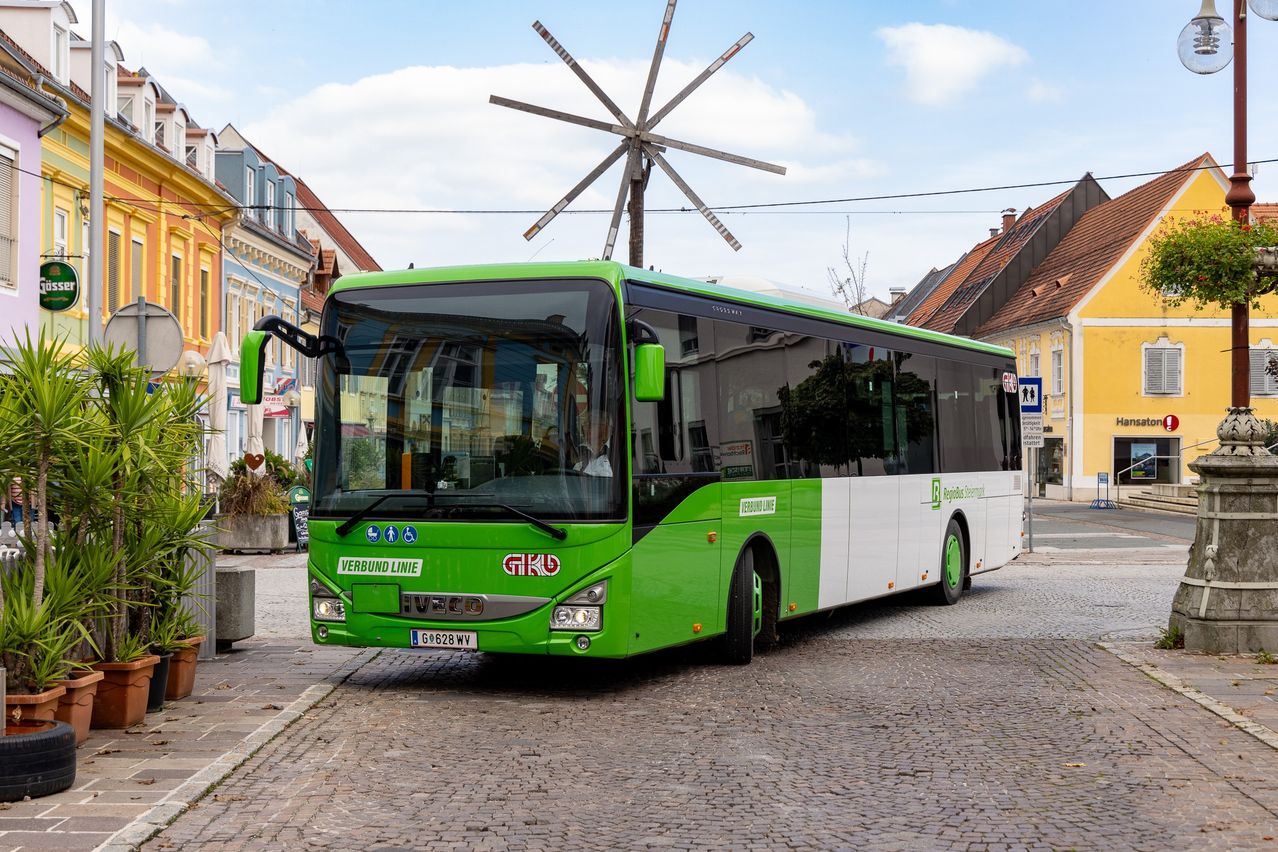 A green and white bus labeled 'Verbund Linie' is parked in a cobblestone street with buildings and a windmill in the background.
