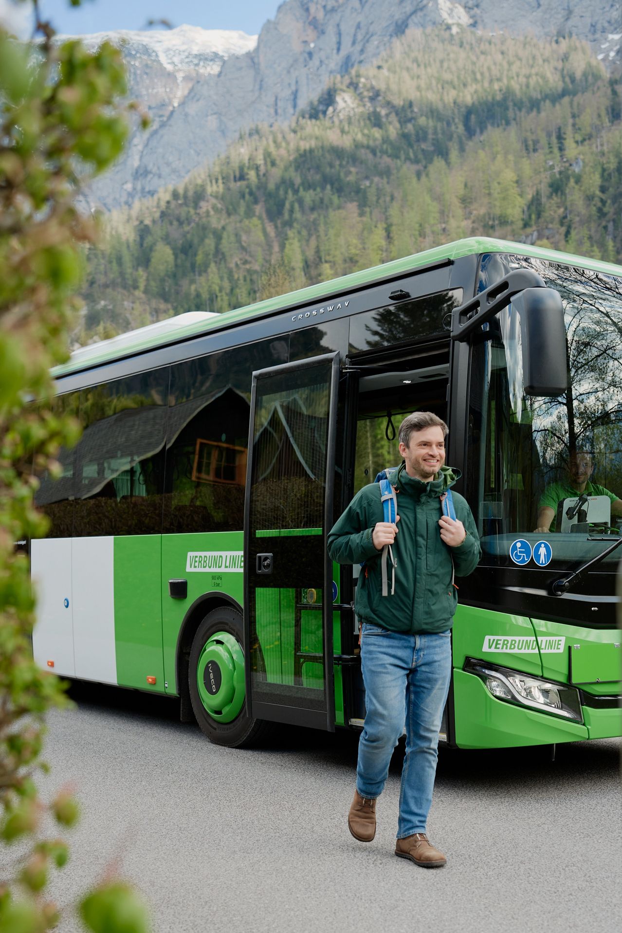 Ein Mann mit einem Rucksack geht auf die offene Tür eines grünen Busses in einem Gebirgsgebiet zu.