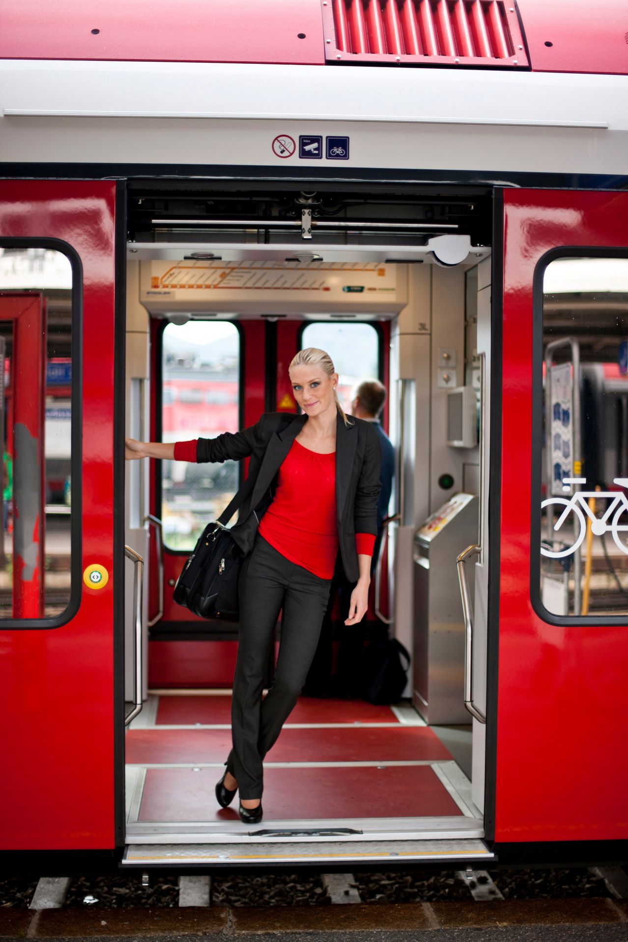 Eine Frau in einem roten Shirt und schwarzem Blazer steigt aus einem roten Zug aus und hält eine schwarze Handtasche. Sie steht vor einer Glastür mit einem Fahrradsymbol.