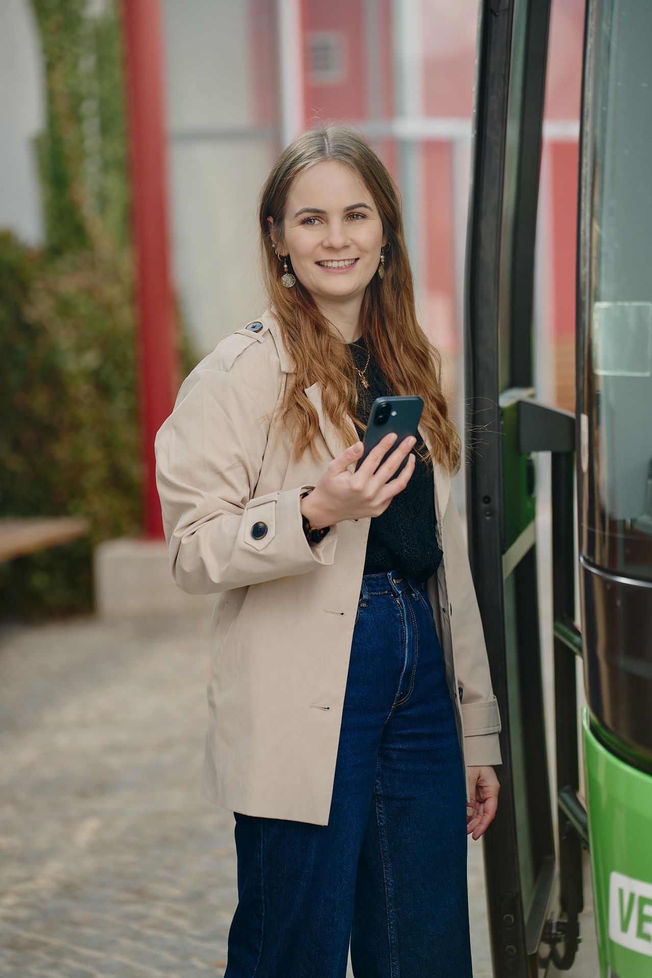 A woman stands outside, holding her phone, smiling, wearing a beige trench coat and blue jeans, near a tram.