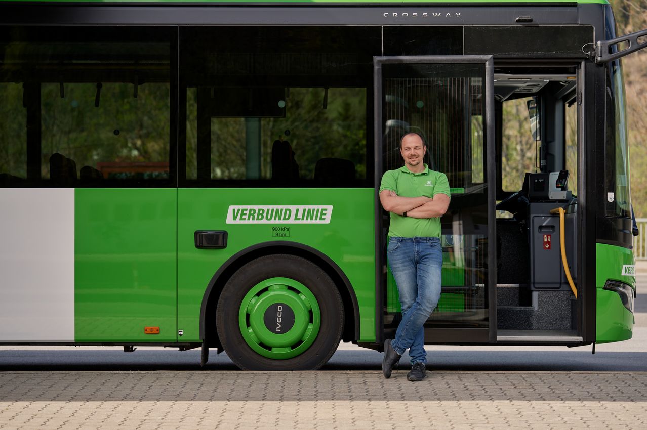 Ein Mann in einem grünen Shirt steht neben einem grünen Bus mit den Worten 'Verbund Linie' an der Seite.