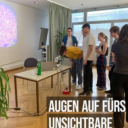 In einem Raum mit einem Mikroskop versammeln sich mehrere Personen. Ein Mann schaut hinein, während andere beobachten. Eine Projektionsleinwand zeigt ein rosa Bild an der Wand.