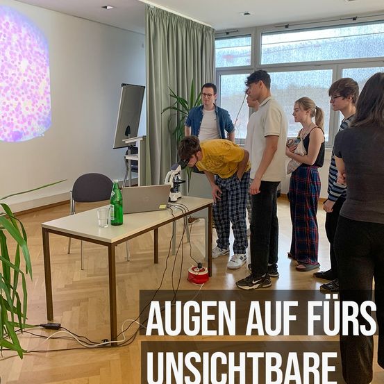 In einem Raum mit einem Mikroskop versammeln sich mehrere Personen. Ein Mann schaut hinein, während andere beobachten. Eine Projektionsleinwand zeigt ein rosa Bild an der Wand.