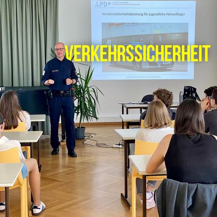 Ein Polizist in Uniform hält eine Verkehrssicherheitsvorlesung für eine Klasse von Studenten, wobei eine Projektionsleinwand eine Sicherheitsbotschaft im Hintergrund anzeigt.