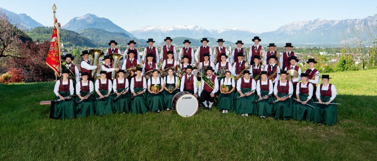 Eine Gruppe von Musikern in traditioneller Kleidung posiert für ein Foto auf einer Rasenfläche mit einer Bergkette im Hintergrund.
