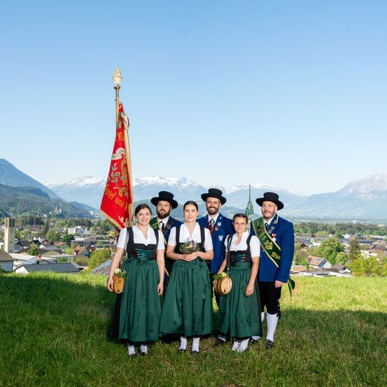 Eine Gruppe von Menschen in traditioneller Kleidung posiert für ein Foto auf einem grasbewachsenen Hügel mit Bergen und einer Stadt im Hintergrund.