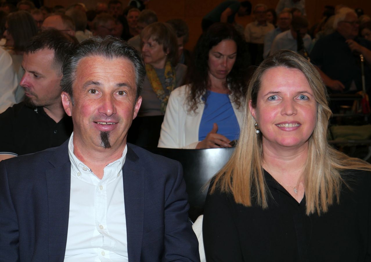 Zwei Personen sitzen in einem Konferenzraum, wobei einer der Männer einen Anzug und einen Schnurrbart trägt und eine Frau ein schwarzes Hemd und eine Halskette trägt. Hinter ihnen sitzen viele Menschen, und einige stehen.