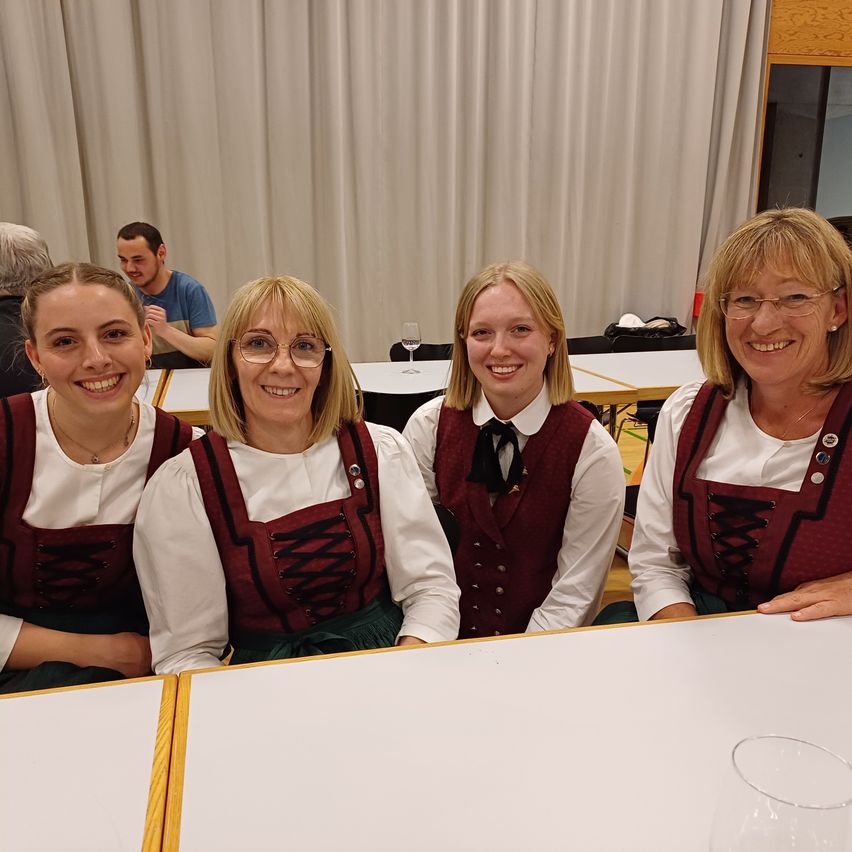 Vier Frauen in traditioneller deutscher Kleidung, lächeln für ein Foto in einem Raum mit weißen Vorhängen und einem Tisch mit Weingläsern.