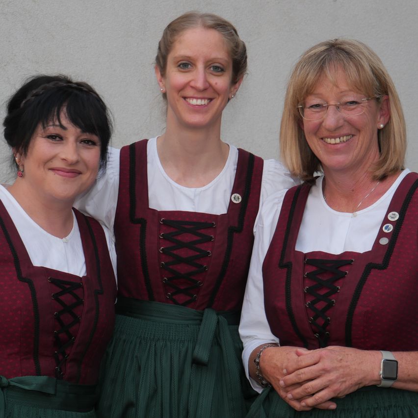 Drei Frauen stehen eng beieinander und tragen traditionelle deutsche Kleidung. Die Frau auf der linken Seite hat ihre Haare geflochten. Die Frau in der Mitte lächelt und die Frau auf der rechten Seite trägt eine Brille.