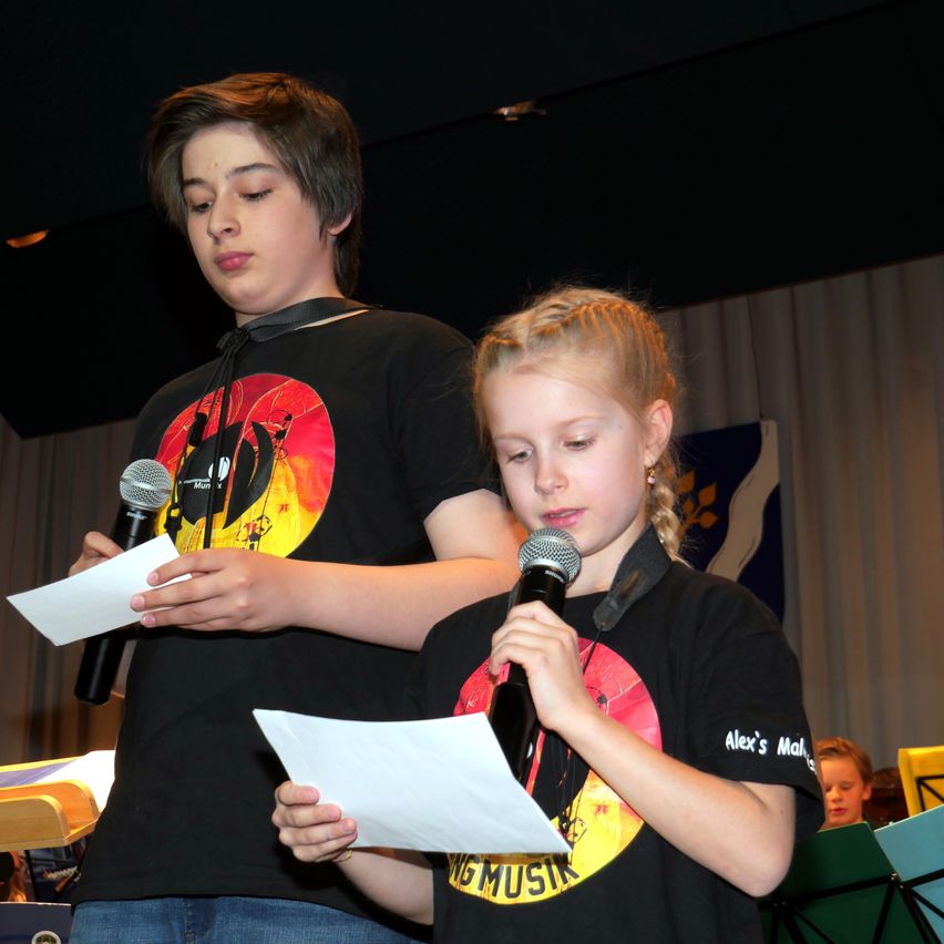 Zwei Kinder stehen auf der Bühne. Der Junge liest von einem Stück Papier und hält ein Mikrofon. Das Mädchen hält ebenfalls ein Mikrofon und hat die Augen geschlossen. Hinter ihnen befindet sich eine Gardine und eine Flagge.