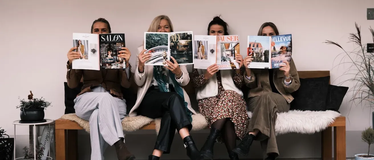 Vier Frauen sitzen auf einer Bank und halten jeweils eine Zeitschrift in der Hand. Sie alle lächeln und schauen in die Kamera. Die Zeitschriften, die sie halten, sind mit 'SALON', 'AD', 'AUSSER' und 'BELLEVUE' beschriftet. Sie sind in lässiger Kleidung gekleidet.