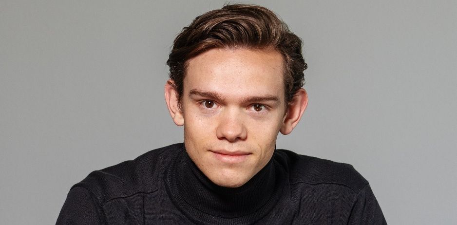 Ein Mann mit kurzen braunen Haaren, der ein schwarzes Rollkragenshirt trägt, posiert für ein Porträt vor einem grauen Hintergrund.