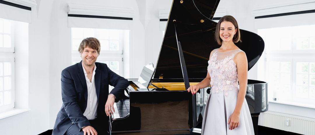 A man and a woman stand beside a grand piano, both smiling and posing for a photo. The man is seated on a piano bench, and the woman stands next to him. The piano is black with gold accents.