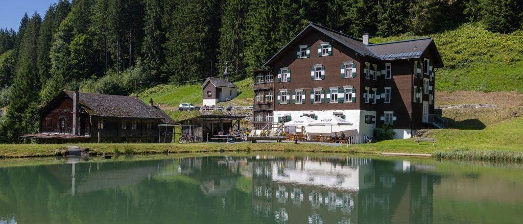 Ein Chalet am Hang mit einem großen See, Bäumen und einer Wasserreflexion. Ein kleineres Gebäude und Autos sind in der Nähe.