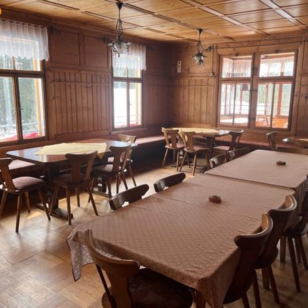 Ein leerer Essbereich mit Holzwänden und Fenstern. Es gibt mehrere Esstische, die mit Tischdecken bedeckt sind, und Holzstühle. Eine Bank steht in der Nähe der Fenster.