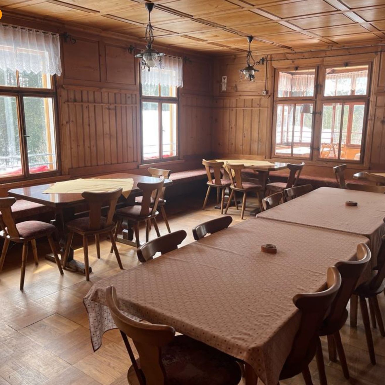 Ein leerer Essbereich mit Holzwänden und Fenstern. Es gibt mehrere Esstische, die mit Tischdecken bedeckt sind, und Holzstühle. Eine Bank steht in der Nähe der Fenster.