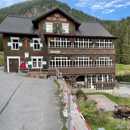 Ein großes braunes Holzgebäude mit grünen Fensterläden und einem roten Dach ist zu sehen, mit Bergen im Hintergrund.