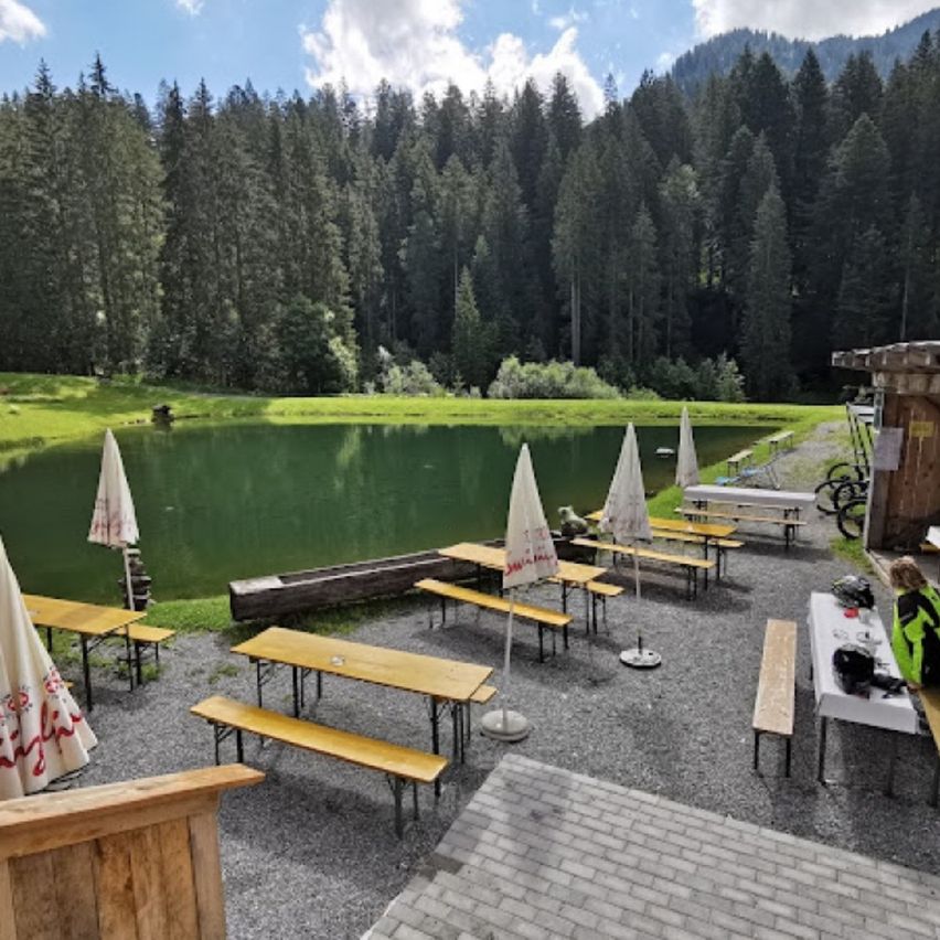 A serene outdoor dining area with wooden benches and tables, surrounded by a lush green landscape and a calm lake, under a partly cloudy sky.