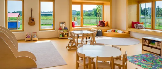 Ein Kinderzimmer mit hellen Wänden, großen Fenstern, einem Tisch mit Stühlen und einer Gitarre, die an der Wand hängt.