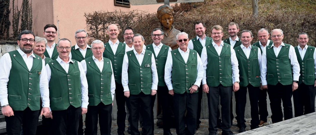 Eine Gruppe von Männern in grünen Westen und schwarzen Hosen steht vor einer Statue, mit einem Ziegelweg und einer Betonmauer im Hintergrund.
