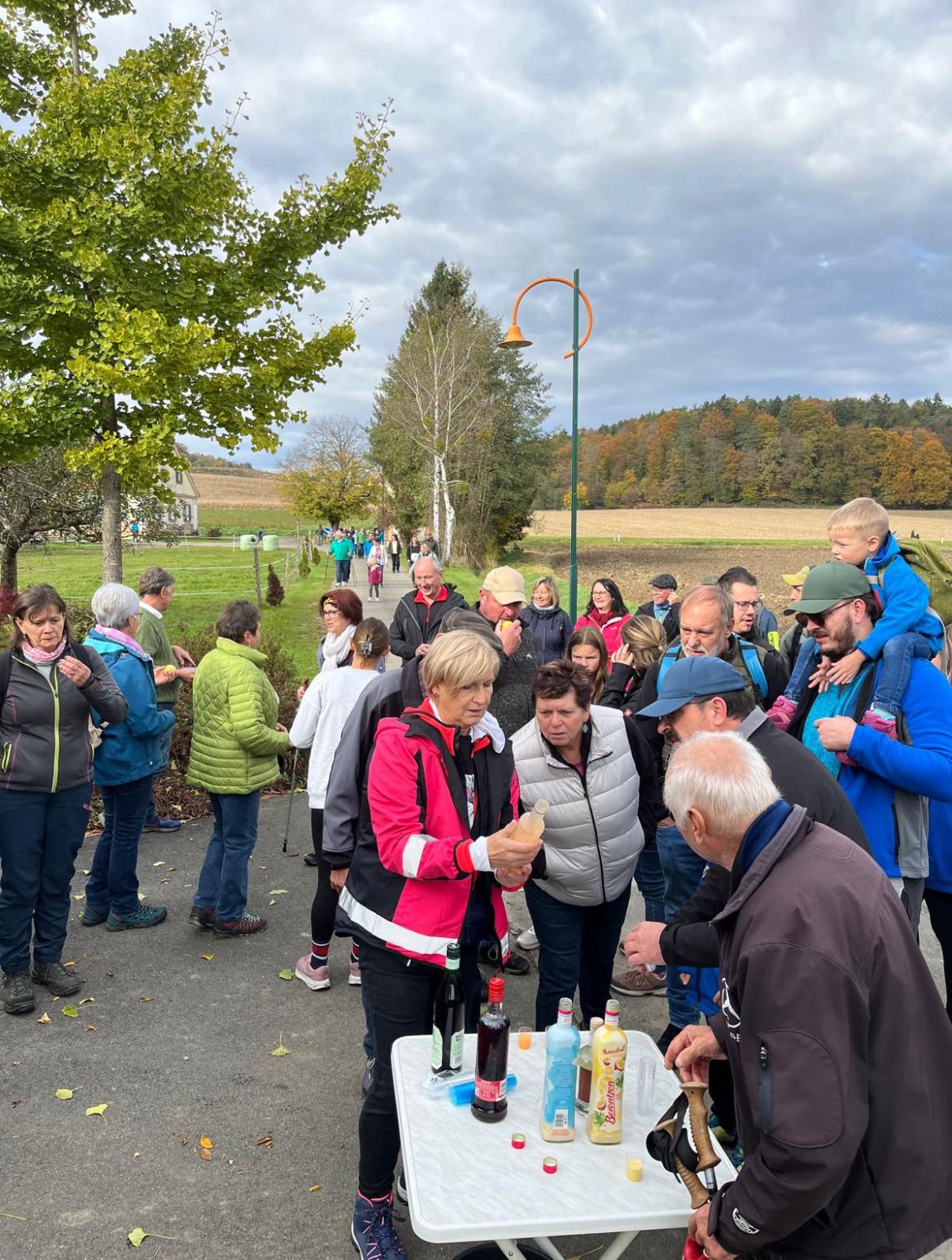 Eine Gruppe von Menschen versammelt sich in der Nähe eines Tisches mit Flaschen Likör. Einige tragen Jacken und Turnschuhe. Bäume und ein Feld sind im Hintergrund.