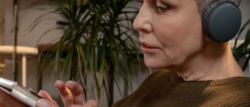 Eine ältere Frau mit grauem Haar sitzt auf einem Sofa, trägt Kopfhörer und hält ein Handy. Sie scheint etwas zu hören. Hinter ihr stehen große Topfpflanzen.