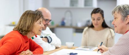 Drei Personen sitzen um einen Tisch in einer Küche. Eine Frau in einem roten Pullover lächelt, während eine andere Frau schreibt. Ein Mann in einem weißen Hemd beobachtet. Auf dem Tisch liegen Utensilien wie Schere, Klebeband und farbige Stifte.