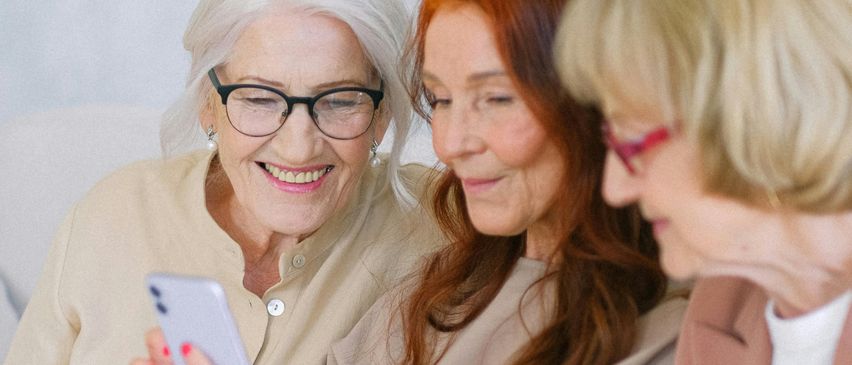 Drei ältere Frauen sitzen zusammen, lächeln und schauen auf ein Smartphone. Die Frau auf der linken Seite hält das Handy, während die anderen zuschauen.
