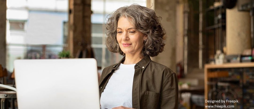 Eine ältere Frau mit grauem Haar sitzt an einem Schreibtisch mit Laptop und Maus und lächelt. Sie trägt ein weißes Shirt und eine braune Jacke. Hinter ihr ist ein großes Fenster.