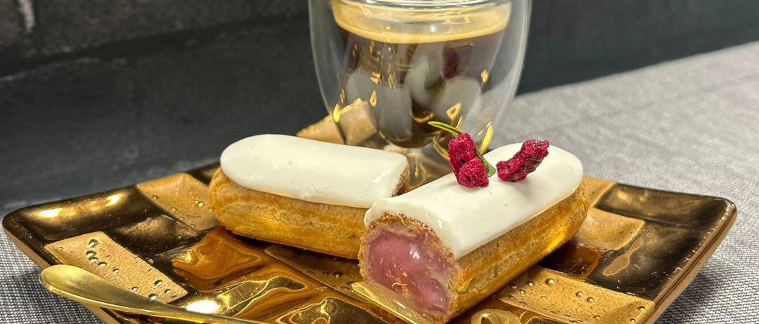 Eine goldene Platte mit zwei Desserts, eines mit weißer Creme und das andere mit roten Beeren. Ein Glas mit Kaffee steht hinter der Platte.