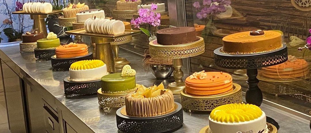 Eine Ausstellung verschiedener Kuchen in verschiedenen Farben und Designs auf einem Tresen, mit einer dekorativen Vase und Blumen im Hintergrund.