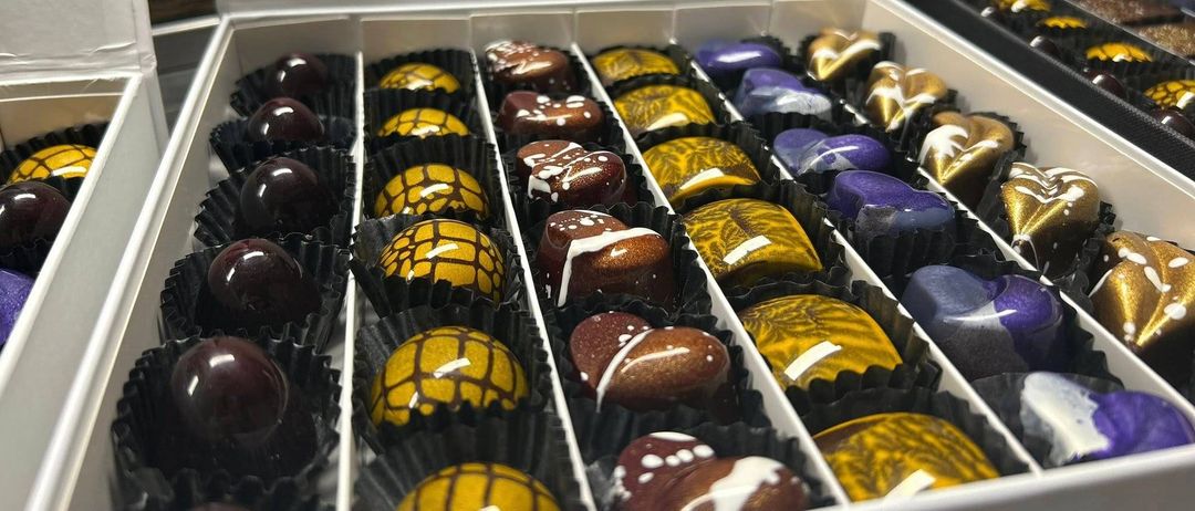 Eine weiße Schachtel gefüllt mit Schokoladen in verschiedenen Farben und Designs, einschließlich Gelb, Lila und Braun, wobei einige mit komplizierten Mustern versehen sind. Die Schokoladen sind in schwarzen und weißen Verpackungen.