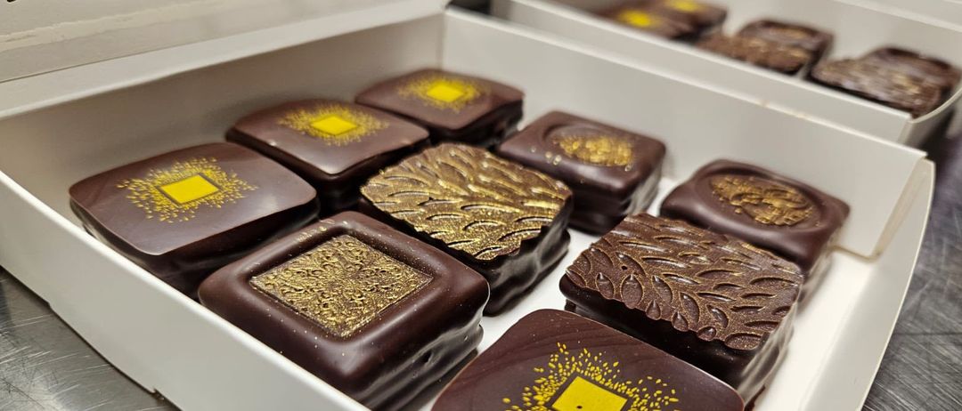 Eine weiße Schachtel gefüllt mit einer Auswahl an Pralinen. Jede Praline hat ein einzigartiges goldenes Design auf der Oberseite und ist in verschiedenen Formen. Die Pralinen sind ordentlich angeordnet.