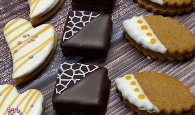 Auf einer Holzoberfläche sind verschiedene Kekse zu sehen. Einige sind herzförmig mit dekorativer weißer Glasur und andere sind mit Schokolade überzogen. Die Kekse variieren in Design und Farbe.