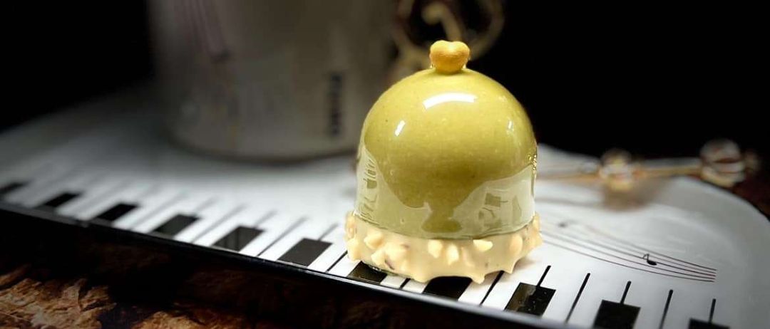 Ein gelbes Dessert in einer Kuppelform steht auf einem Teller mit Klaviertasten-Design. Dahinter ist ein weißer Becher mit einem Griff zu sehen.