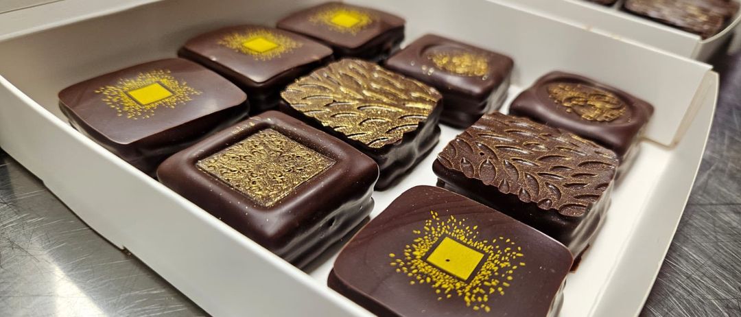 Eine weiße Schachtel enthält mehrere Pralinen mit komplizierten Verzierungen auf einer glänzenden Oberfläche, die auf einem Edelstahltisch platziert sind.