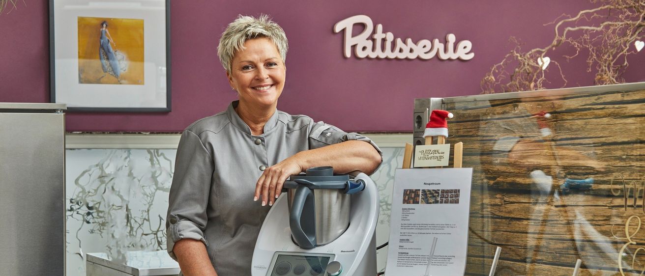 Eine Frau steht lächelnd hinter einer Theke in einer Patisserie. Sie hat eine Hand auf einem weißen Gerät, wahrscheinlich einem Mixer. Hinter ihr ist eine Wand mit dem Wort 'Patisserie'. Auf dem Tresen sind zwei Honigflaschen, ein Tuch und ein Schild. Ein Bilderrahmen ist links.