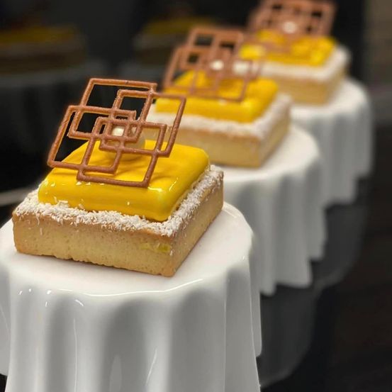 Fünf Zitronenkuchen mit dekorativen braunen Quadraten obenauf stehen auf weißen Ständern. Die Kuchen sind mit gelber Creme und Kokosnuss garniert.