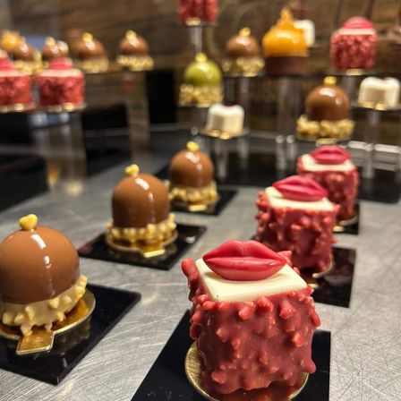 Ein Tisch ist mit verschiedenen Schokoladen-Desserts gefüllt. Es gibt Schokoladen-überzogene Kuchen mit Toppings, und einige haben rote Lippenstift-förmige Dekorationen. Im Hintergrund befinden sich weitere Desserts auf Ständern, darunter welche mit roter, gelber und grüner Glasur.