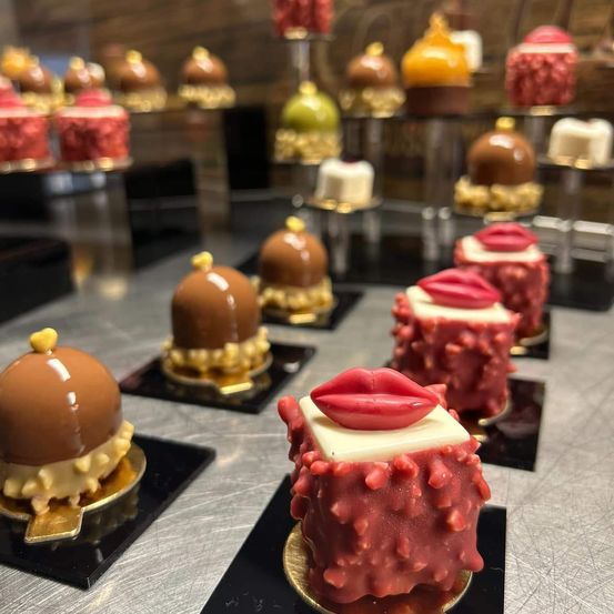 Ein Tisch ist mit verschiedenen Schokoladen-Desserts gefüllt. Es gibt Schokoladen-überzogene Kuchen mit Toppings, und einige haben rote Lippenstift-förmige Dekorationen. Im Hintergrund befinden sich weitere Desserts auf Ständern, darunter welche mit roter, gelber und grüner Glasur.