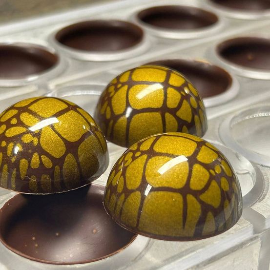 Drei goldene und braune Schokoladentrüffel mit komplizierten Mustern liegen in einer Schokoladenform, umgeben von leeren Fächern.