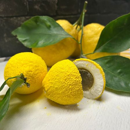 Drei Zitronen mit gelber Schale, zwei davon ganz und eine aufgeschnitten auf einer weißen Oberfläche. Die Blätter sind grün.