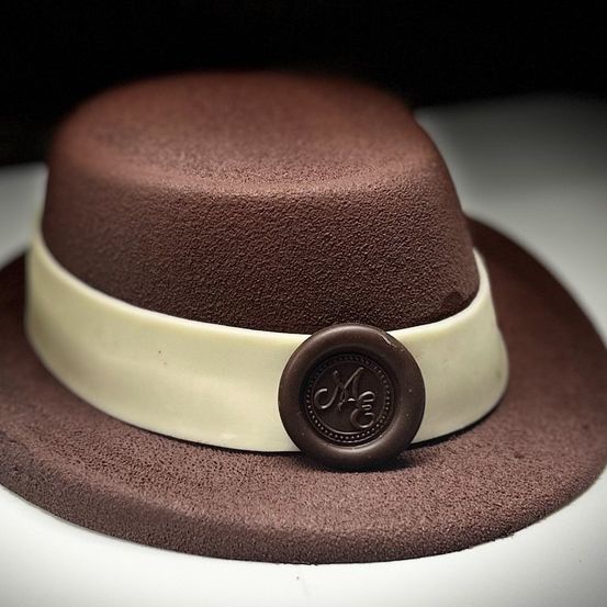 Ein Schokoladen-Fedorahut mit einem braunen Band und einem weißen Cremeband mit einem Schokoladen-Monogrammsiegel darauf.