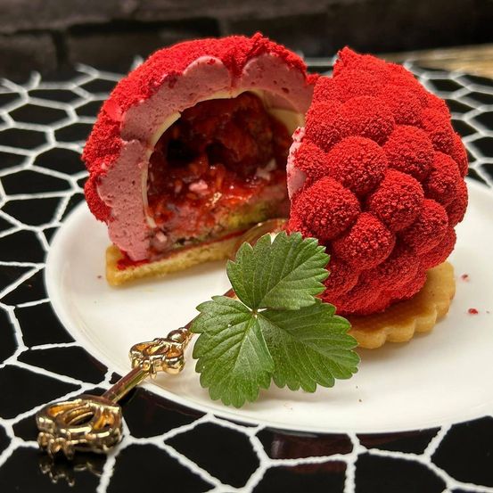 Ein Dessert mit roter Himbeerfüllung, einem Erdbeerblatt und einem goldenen Schlüssel auf einem gemusterten Teller.