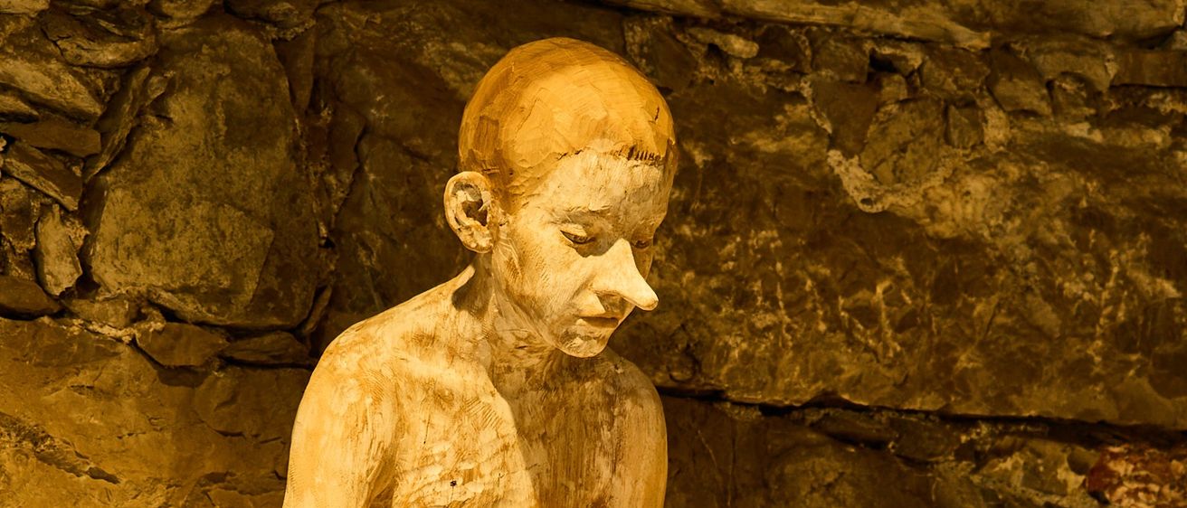 Eine Holzstatue eines Jungen mit dunklem Haar sitzt auf einer Steinmauer. Er ist nackt, und seine Füße sind schwarz bemalt.