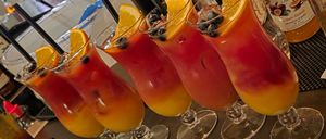 Vier Gläser, gefüllt mit einem leuchtend roten und gelben Getränk, garniert mit Orangenscheiben und Blaubeeren, auf einem Tresen mit Flaschen im Hintergrund.