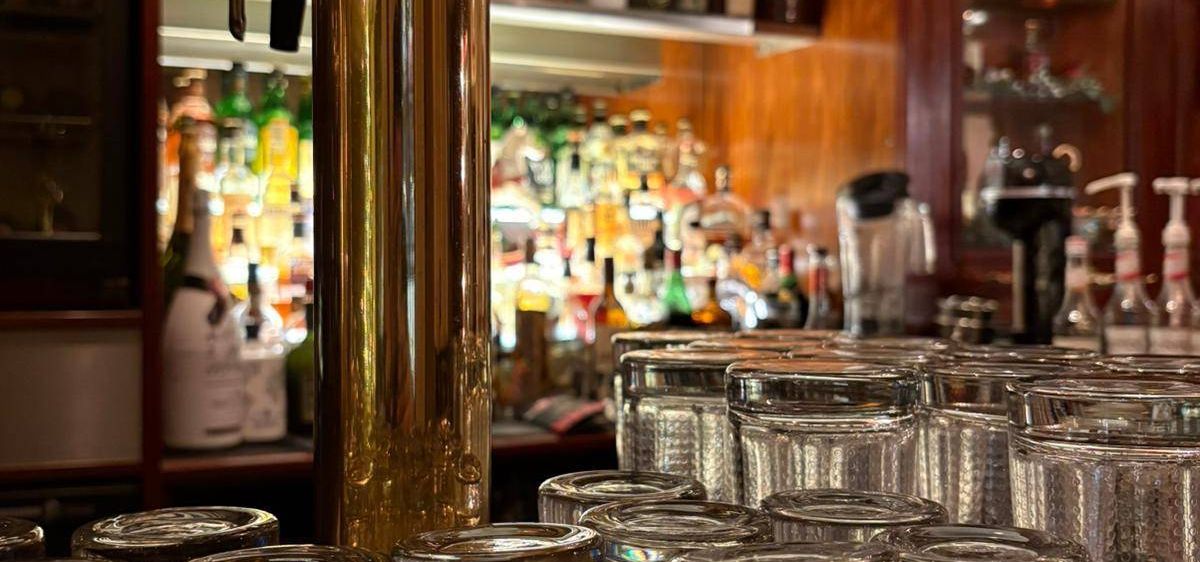 Eine Bar mit Holzregalen und einem goldenen Zapfhahn in der Mitte, gefüllt mit ordentlich angeordneten Flaschen und Gläsern. Im Vordergrund hält ein Tablett mehrere klare Gläser.