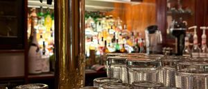 Eine Bar mit Holzregalen und einem goldenen Zapfhahn in der Mitte, gefüllt mit ordentlich angeordneten Flaschen und Gläsern. Im Vordergrund hält ein Tablett mehrere klare Gläser.