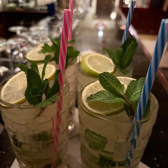 Drei Gläser Mojitos mit Minzblättern und Zitronenscheiben, garniert mit rosa und blau gestreiften Strohhalmen, stehen auf einer Bartheke.
